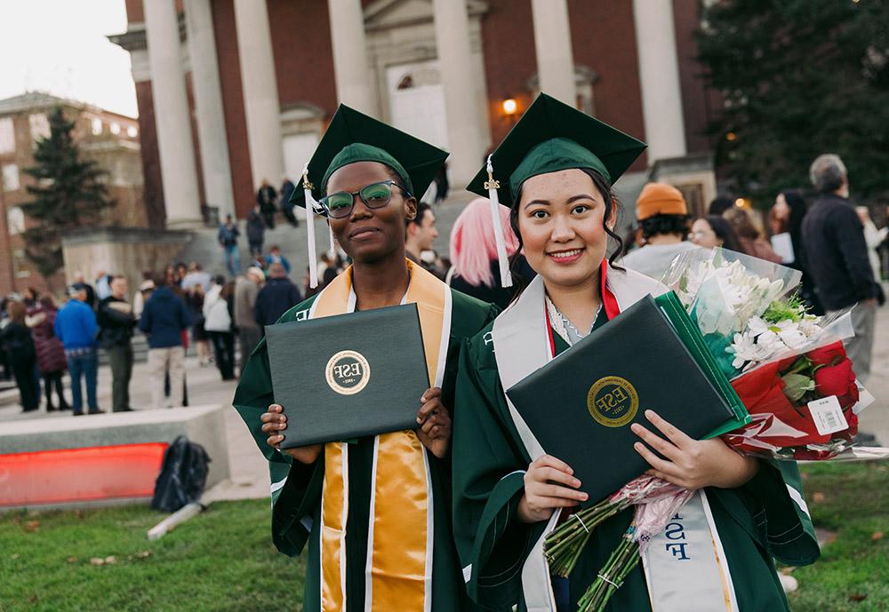 Two female students holding diplomas outside a university building