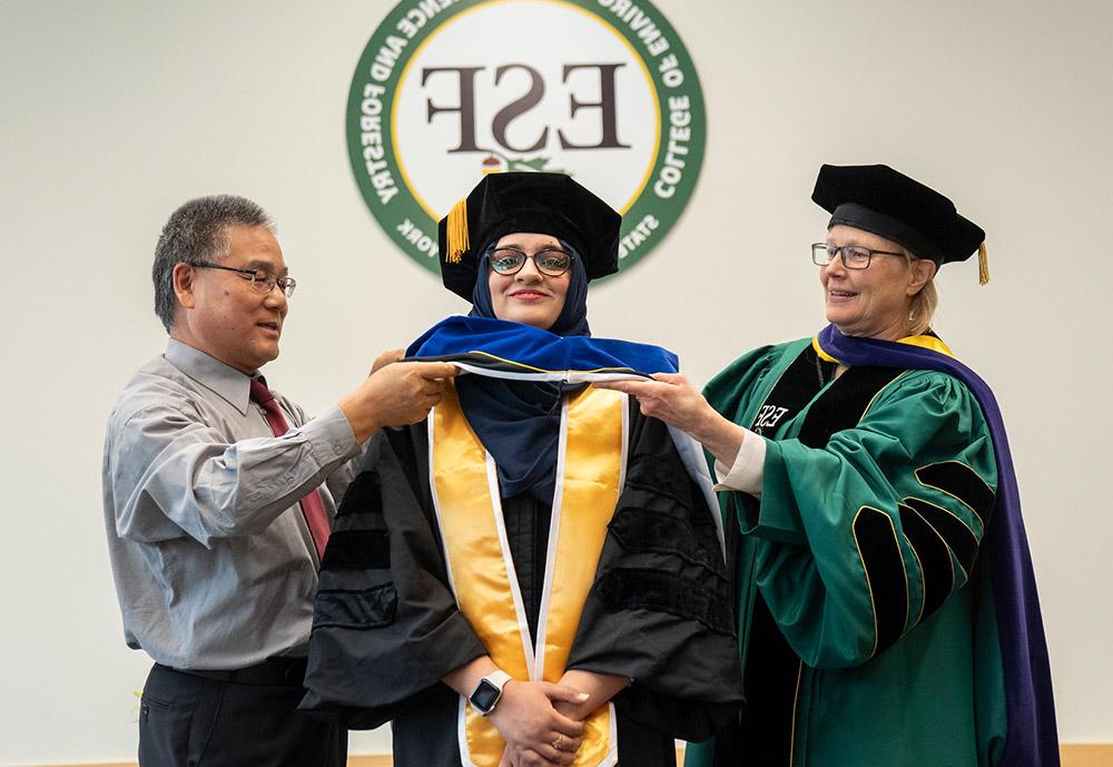 Female students being hooded by college president Joanie Mahoney (left) and male professor (right).